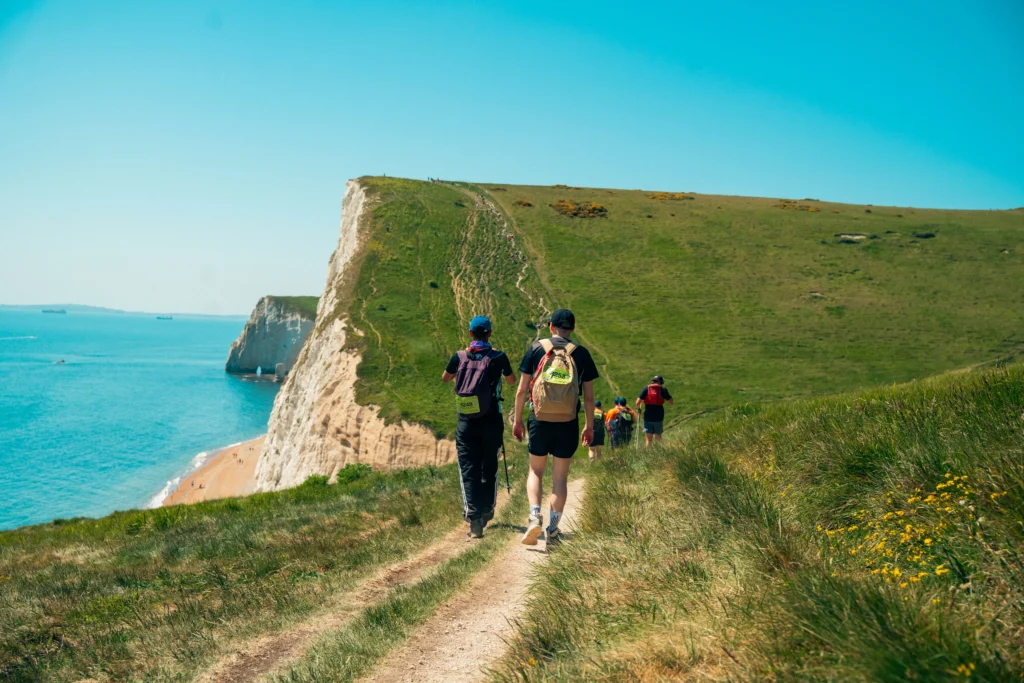People walking along the Jurassic Coast taking part in the Jurassic Coast Ultra Challenge, cliffs in the distance and a bright blue sky.